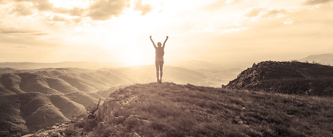 A person standing triumphantly, silhouetted by the sun, having climbed a mountain